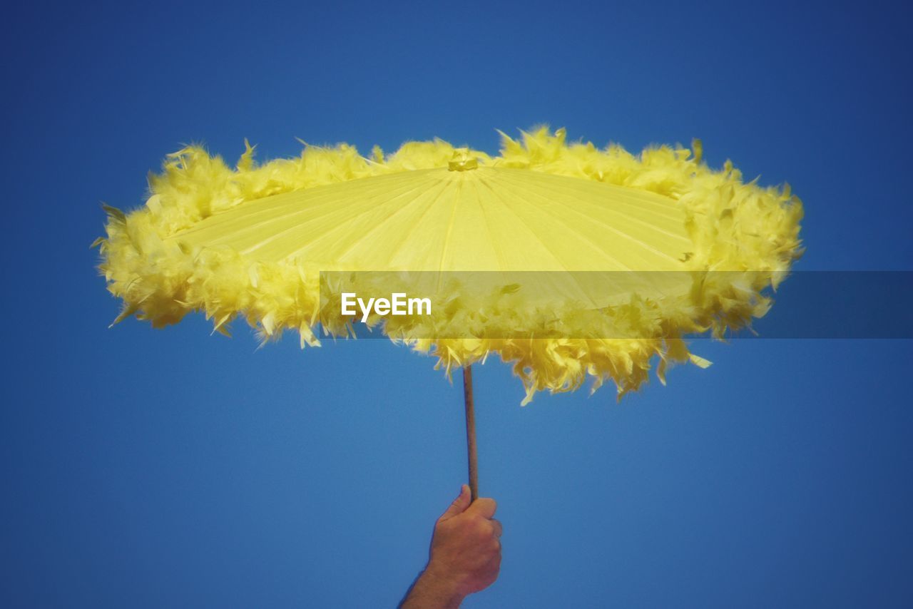 Cropped hand of man holding yellow umbrella against clear blue sky