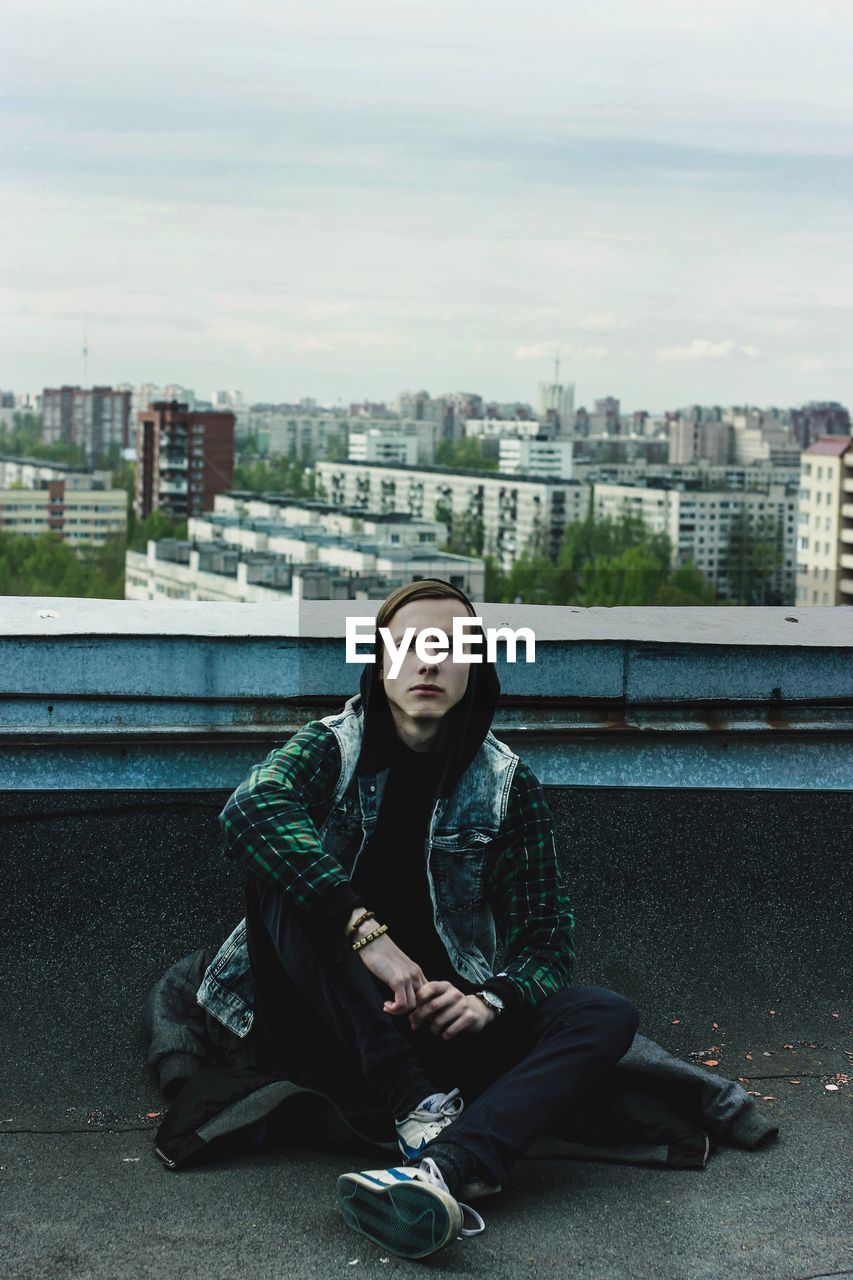 Portrait of man sitting on building terrace against cloudy sky