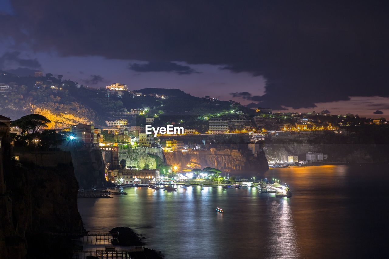 Illuminated city by sea against sky at night