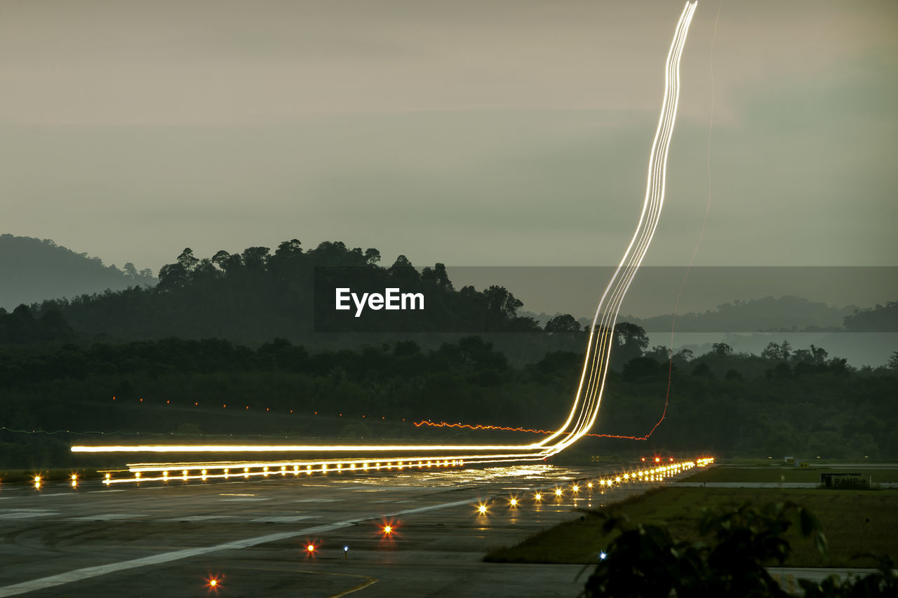 Light trails on road by trees against sky at night