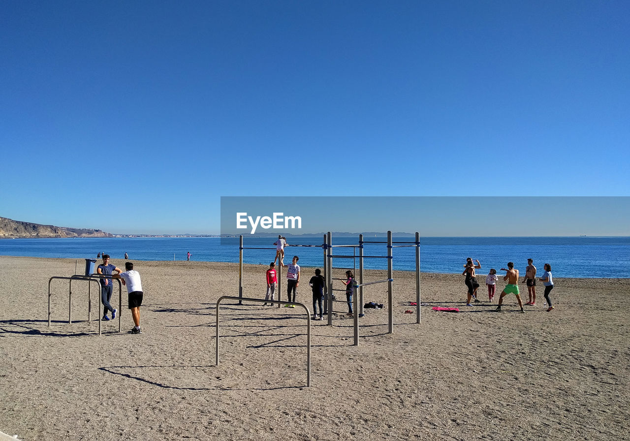 People by play equipment at beach against clear blue sky on sunny day