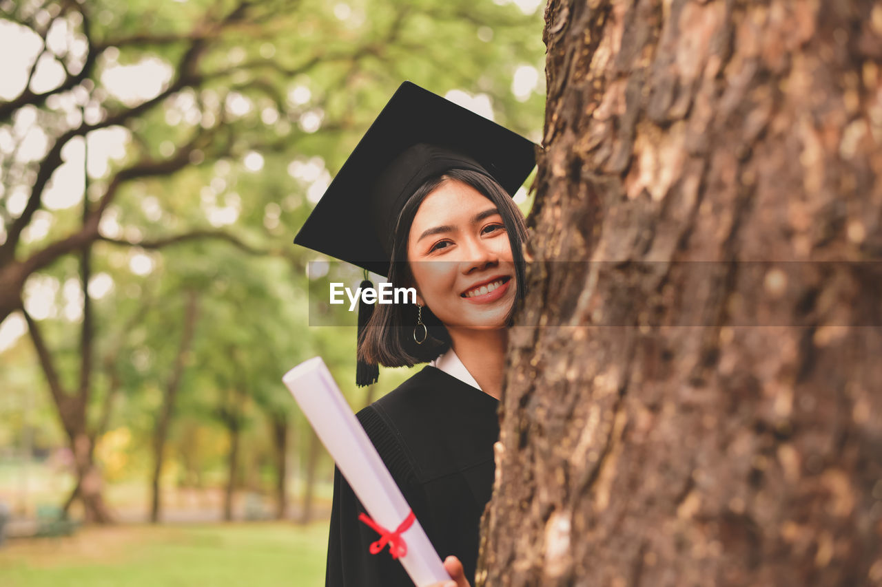 Portrait of young woman in graduation gown holding certificate while standing by tree at park
