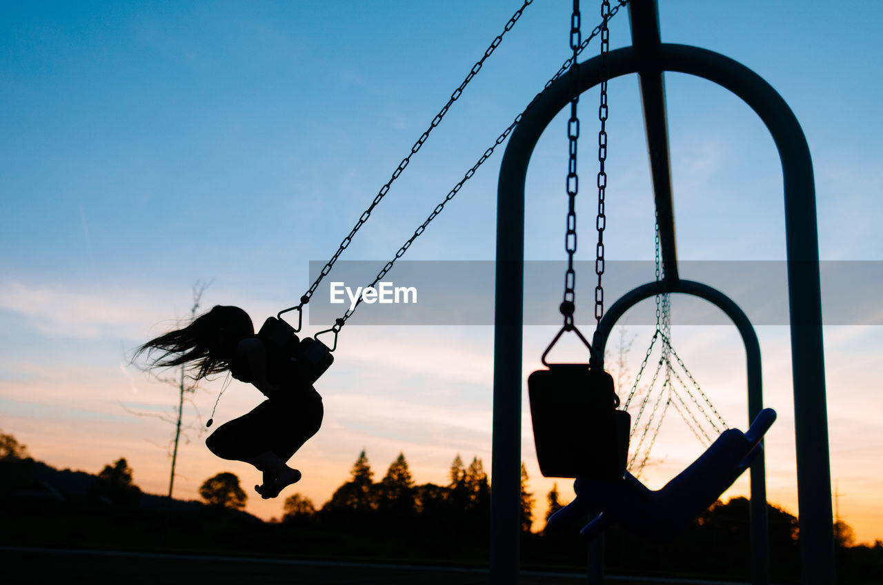 Silhouette girl swinging at playground against sky during dusk