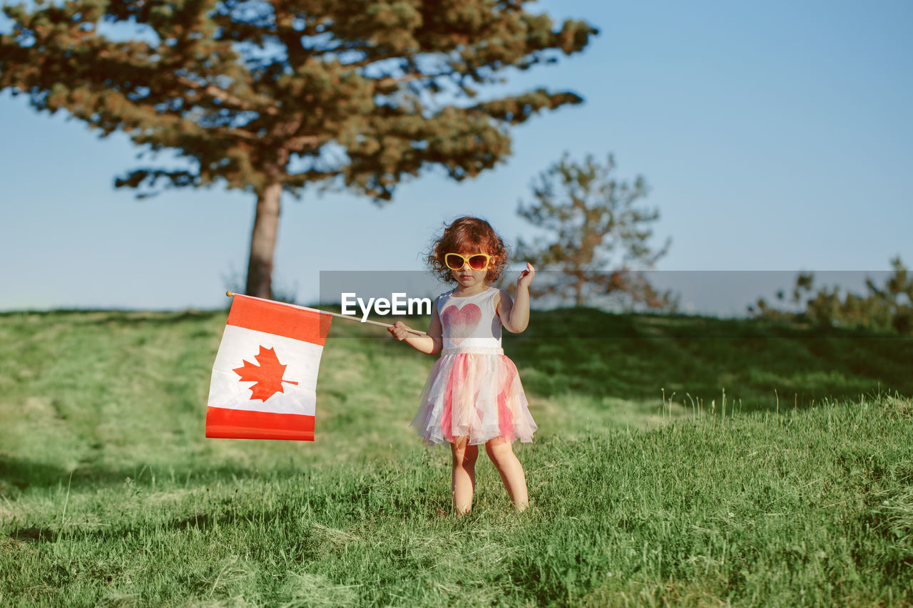 Cute girl holding canadian flag while standing on grass against trees and sky