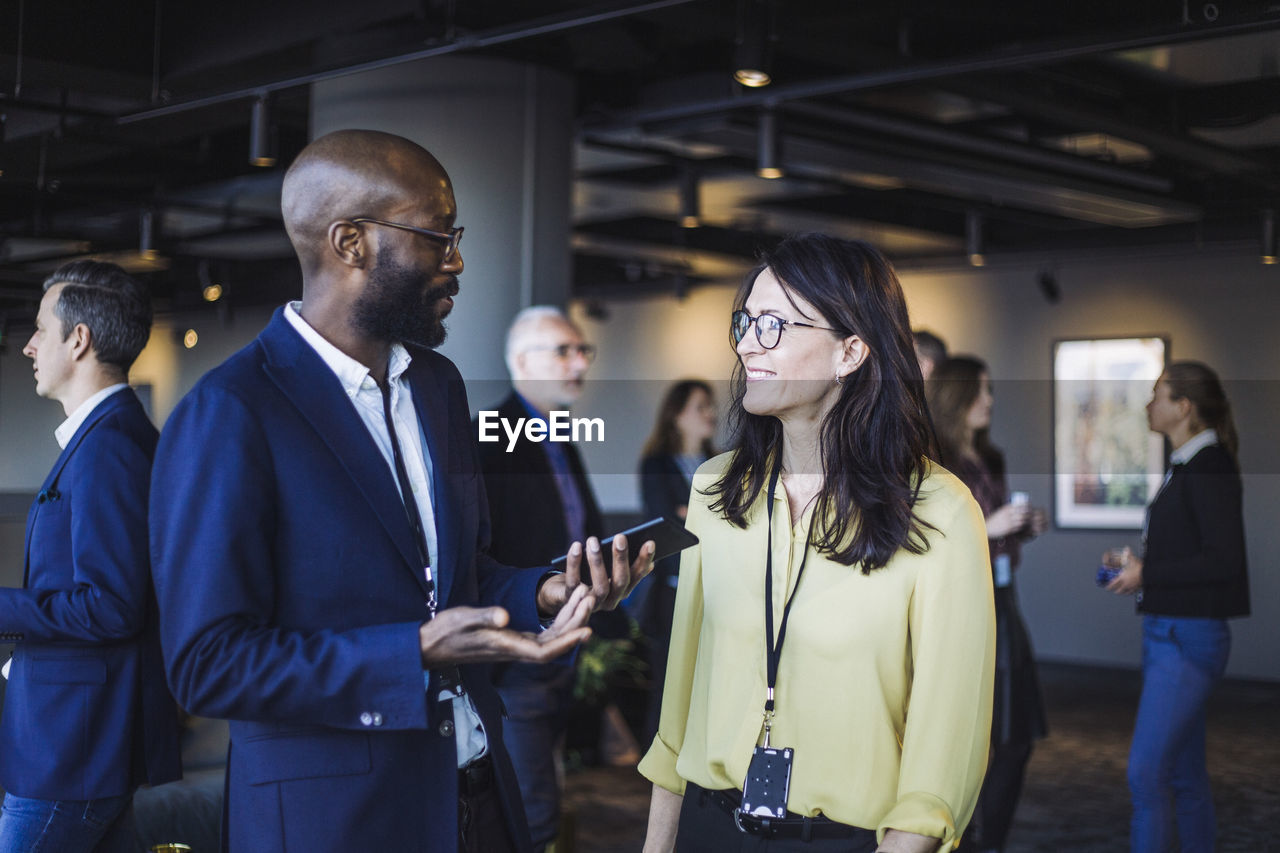 Side view of smiling male with smart phone talking to female colleague in office