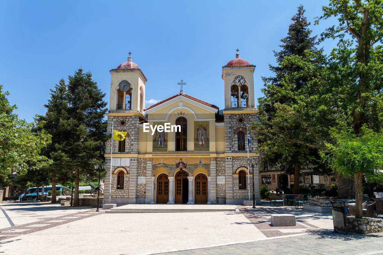 architecture, built structure, building exterior, tree, religion, plant, place of worship, building, nature, sky, belief, travel destinations, estate, spirituality, sunny, blue, history, clear sky, the past, city, sunlight, facade, no people, travel, day, outdoors, landmark, tourism, arch, town square, catholicism