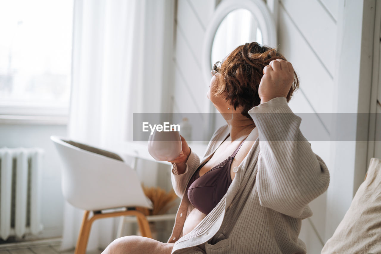 Middle aged plus size woman with brunette curly hair with cup of tea in hand sitting on bed at home