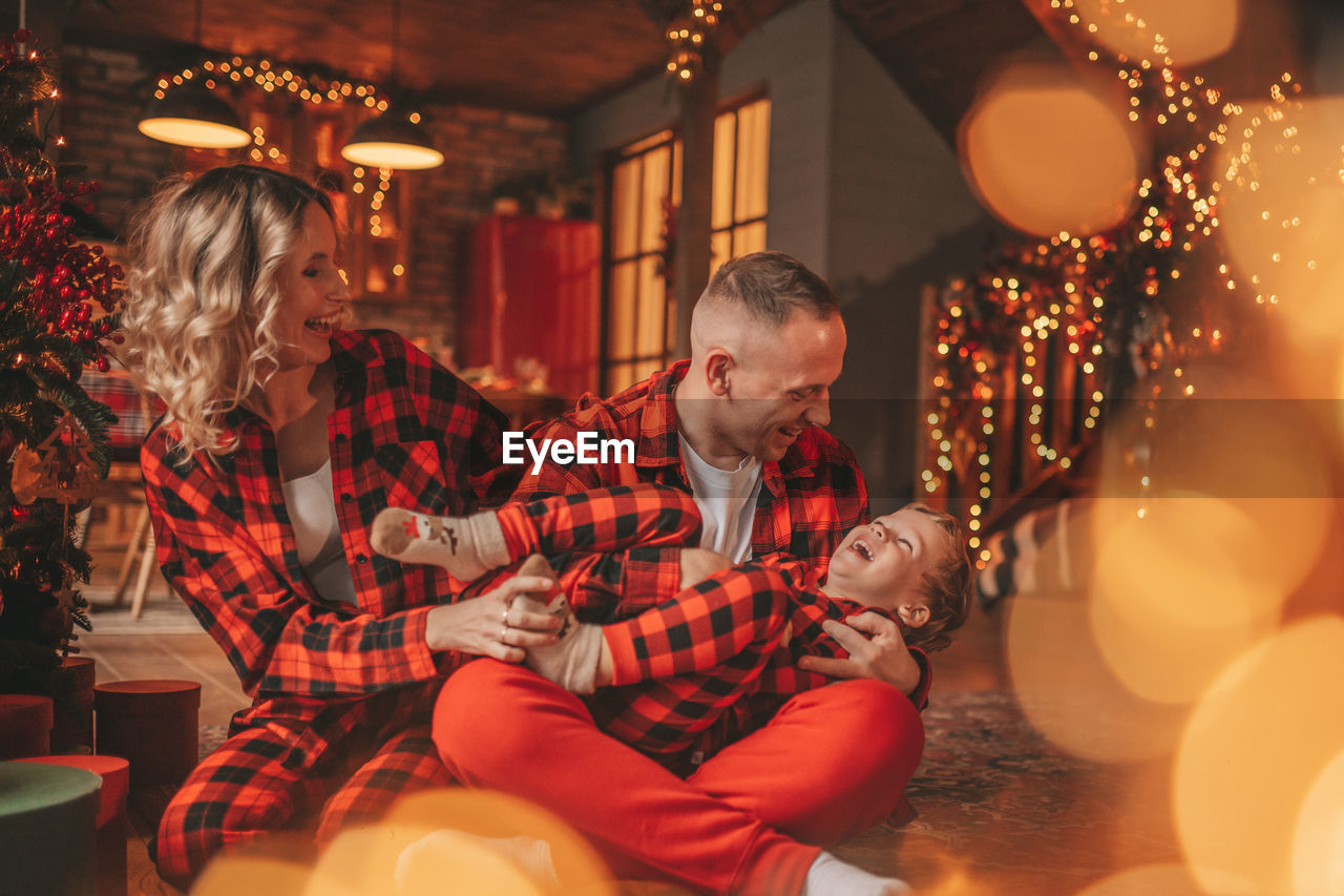 Smile active parents with small son in red checkered sleepwears waiting santa indoor.