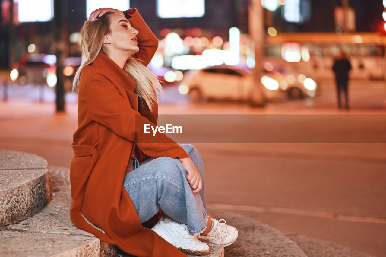 Young woman looking away while sitting on street in city at night