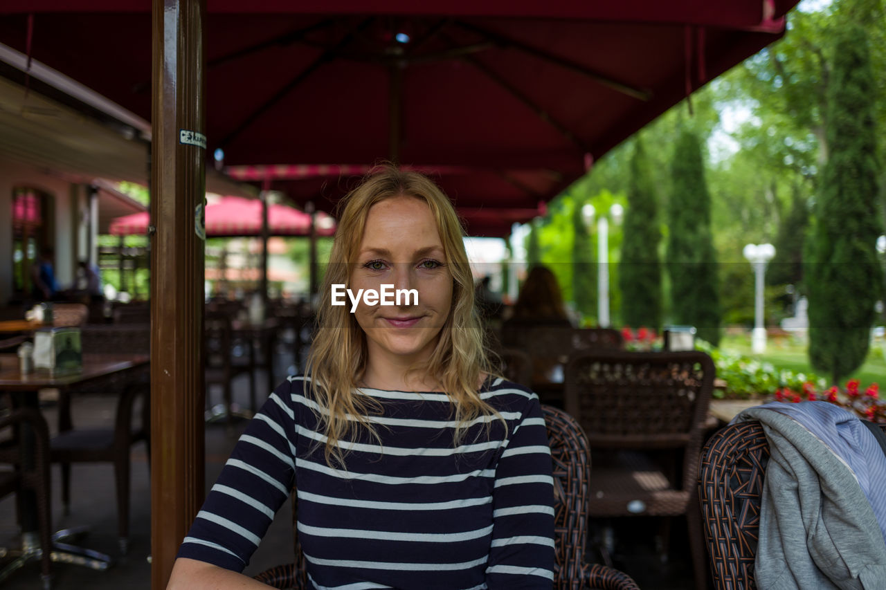 PORTRAIT OF SMILING YOUNG WOMAN STANDING AT OUTDOOR CAFE