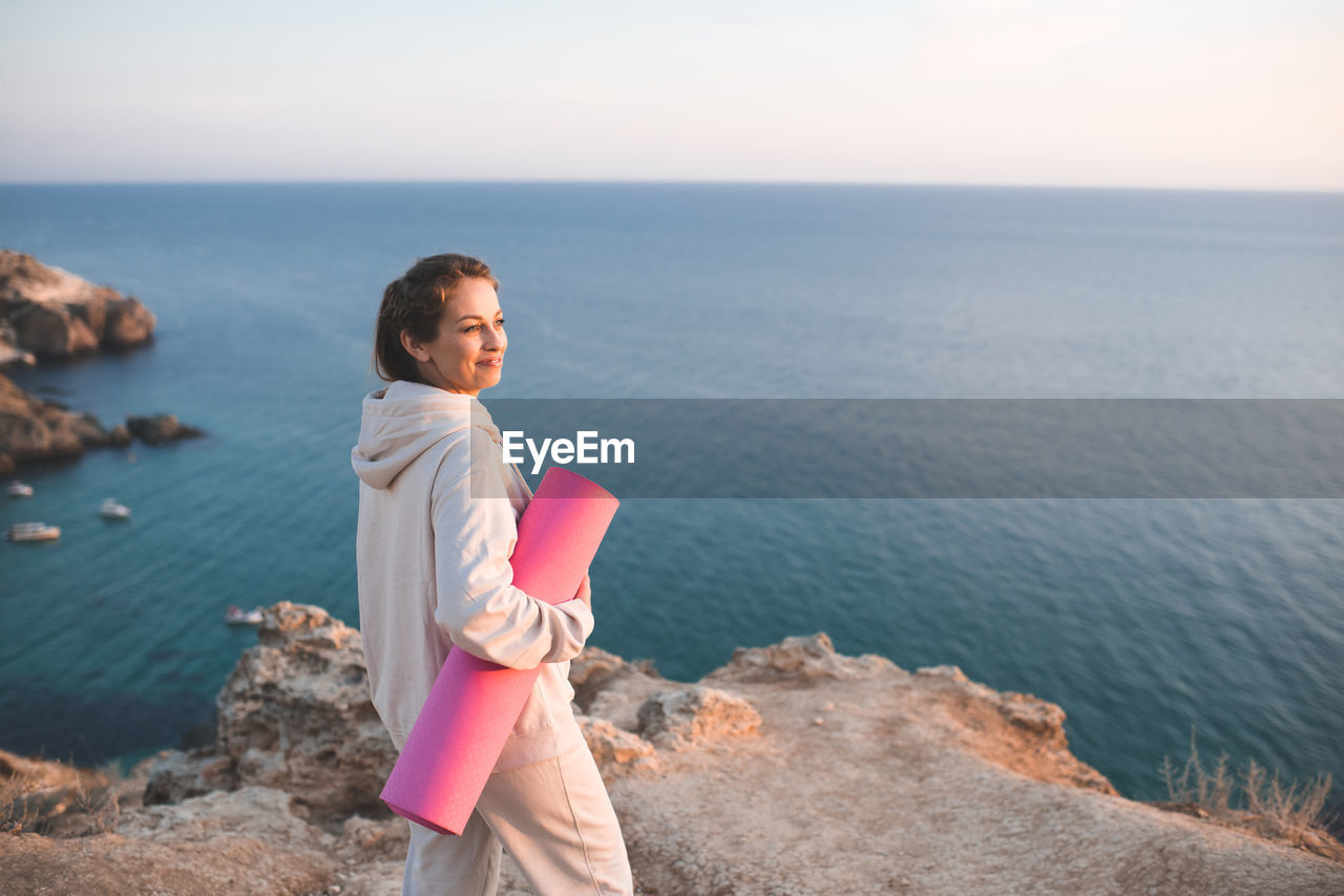 Smiling woman hold yoga mat wear sportswear hoodie and pants stand on top of rock over sea