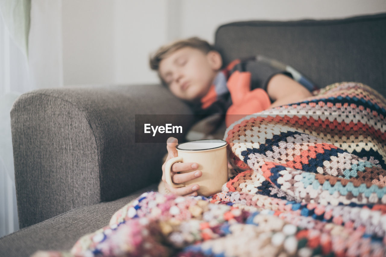 Boy sleeping on sofa with blanket while holding coffee cup in hand at home