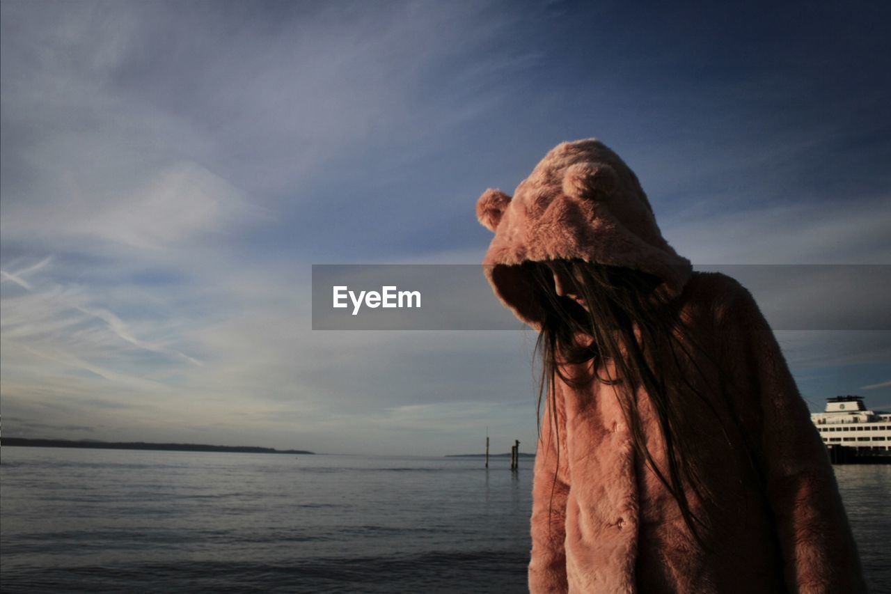 Young woman wearing hooded jacket while standing by sea against sky