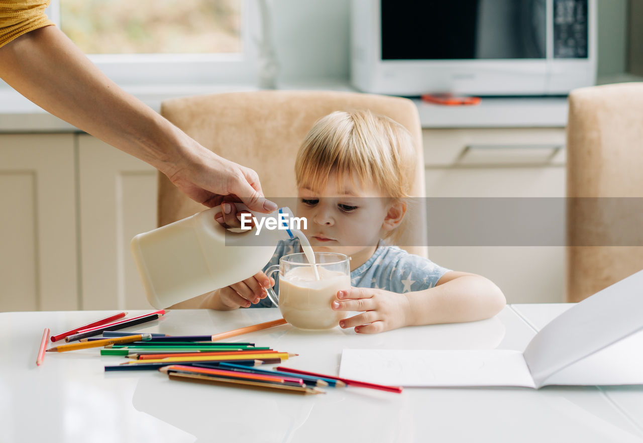 A little girl has breakfast in the kitchen and draws, her mother pours milk into a mug.