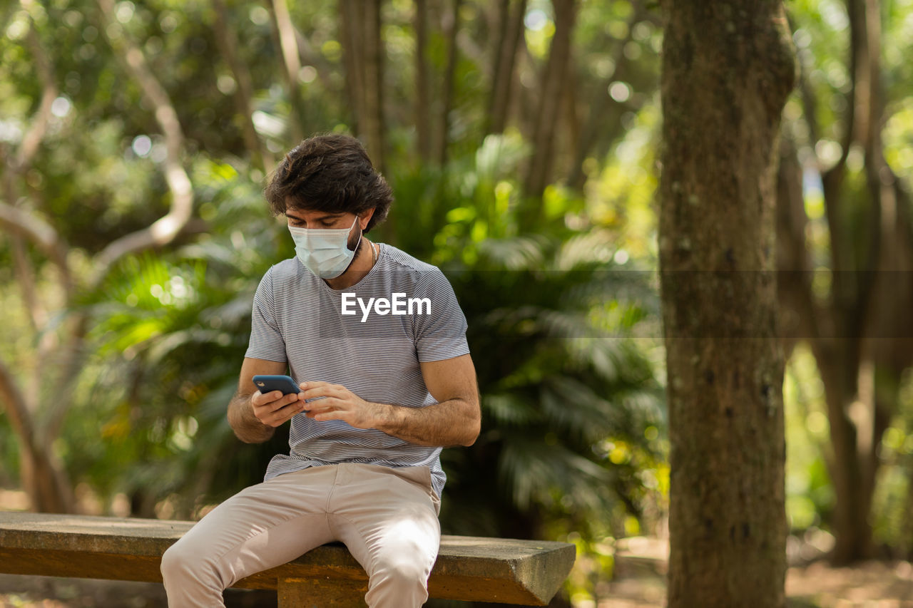 Young man wearing a mask and a smartphone at day time at a green park . mobile phone, technology