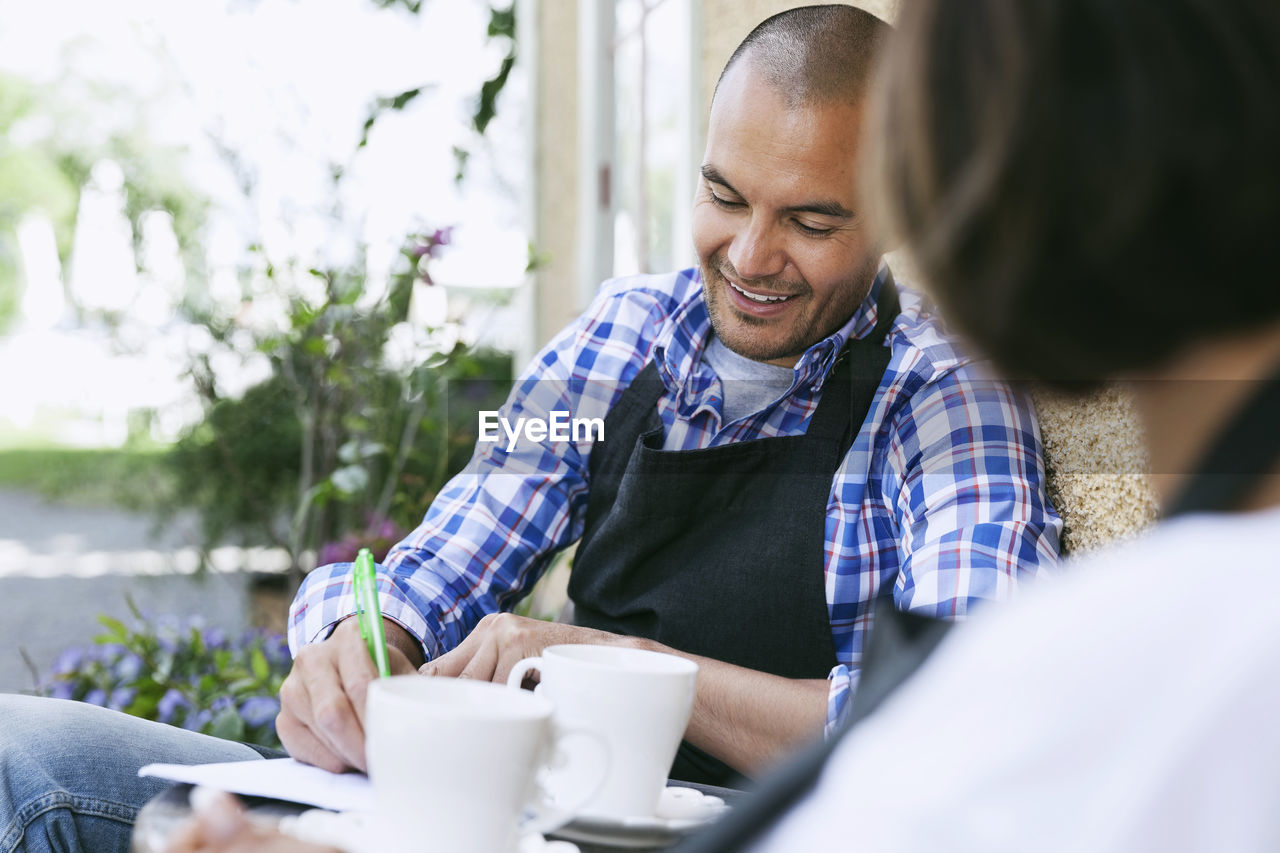 Smiling man writing on paper while having coffee with colleague outside cafe