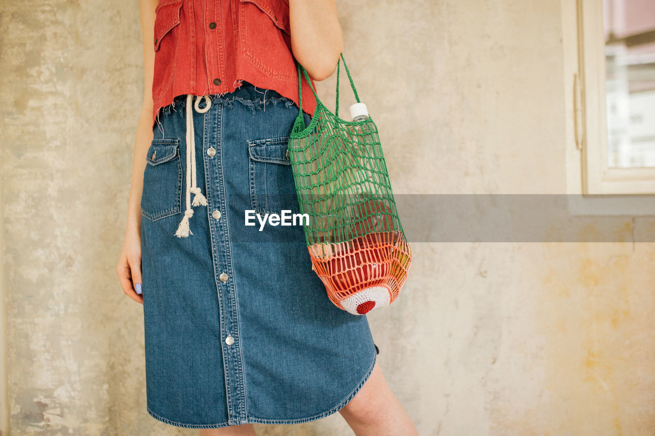 Midsection of woman holding bag standing against wall