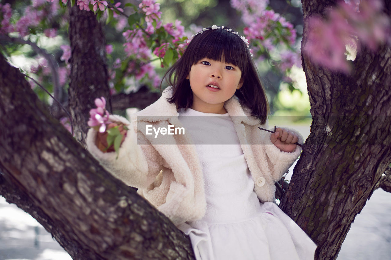 Korean girl in a white light fur coat and a headband sitting on a tree branch in a garden