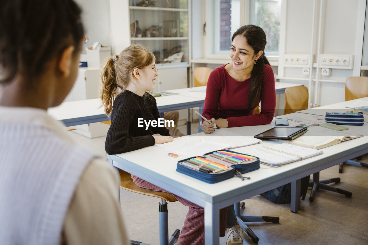 Smiling female teacher and schoolgirl interacting with each other while sitting in classroom