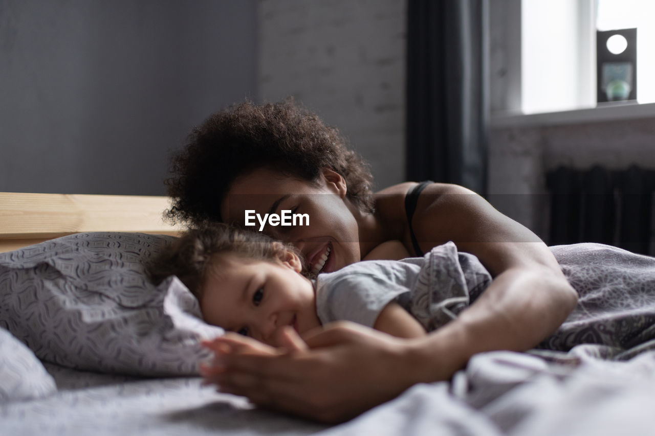 Cheerful mixed race mother and daughter lying on bed in morning