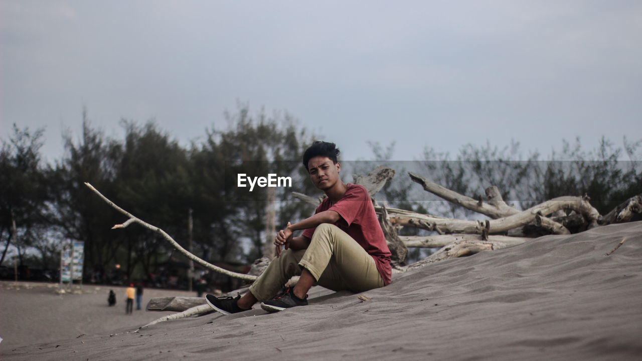 Portrait of young man sitting on sand against sky