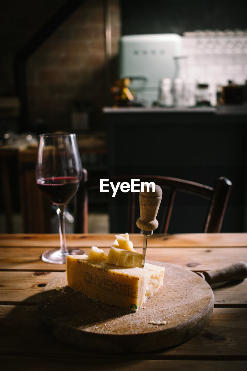 Piece of delicious cheese with knife served on wooden board placed near glass of red wine on wooden plank table in rustic bar