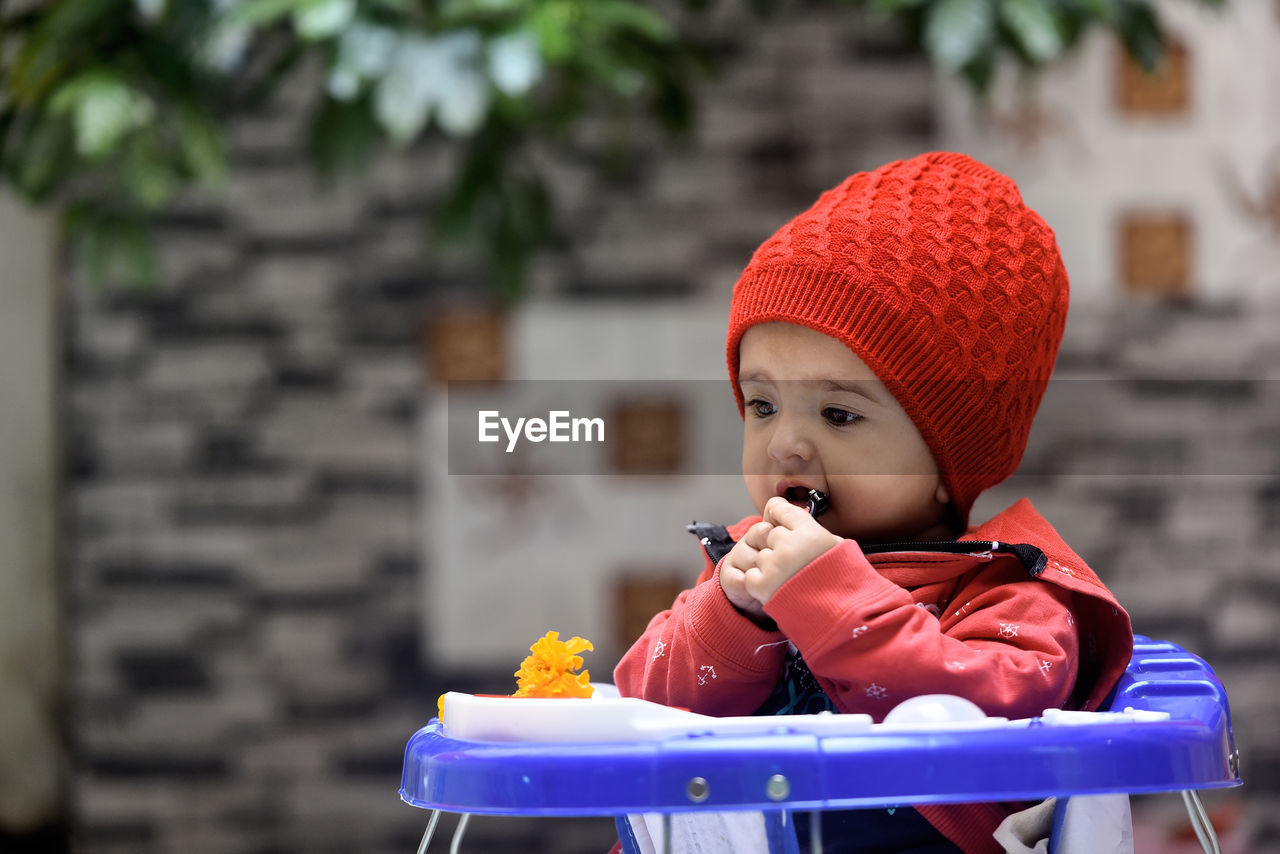 Indian baby girl wearing red winter jacket and woolen cap sitting on a blue walker 