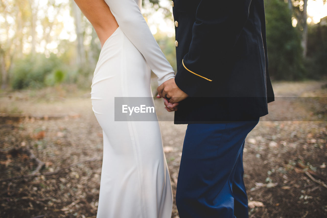 Midsection of bride and groom holding hands and standing on field