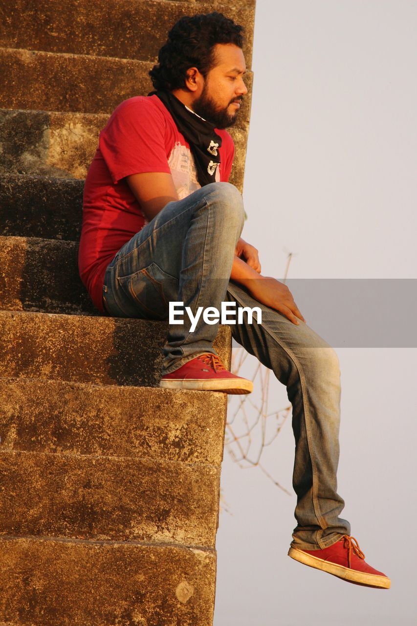 YOUNG MAN USING MOBILE PHONE WHILE SITTING ON WALL