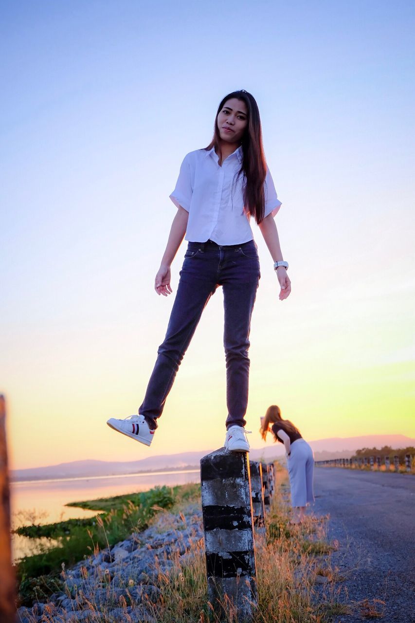 Full length portrait of smiling woman standing on bollard against sky during sunset