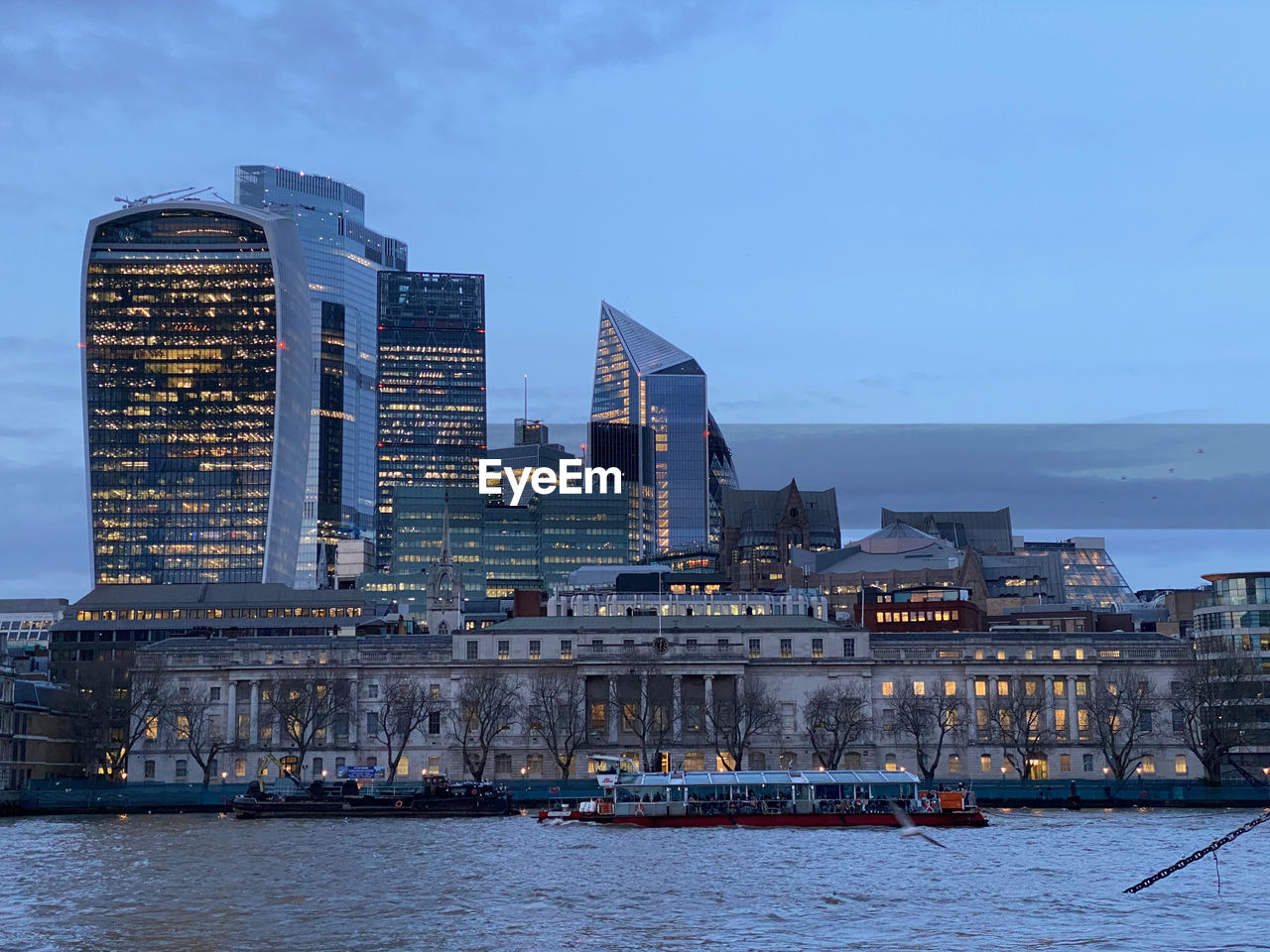 Modern buildings in city of london by river thames against sky at dusk