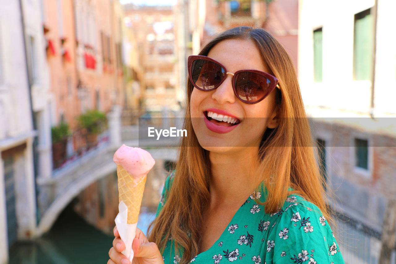 Portrait of cheerful young woman wearing sunglasses while having ice cream cone in city