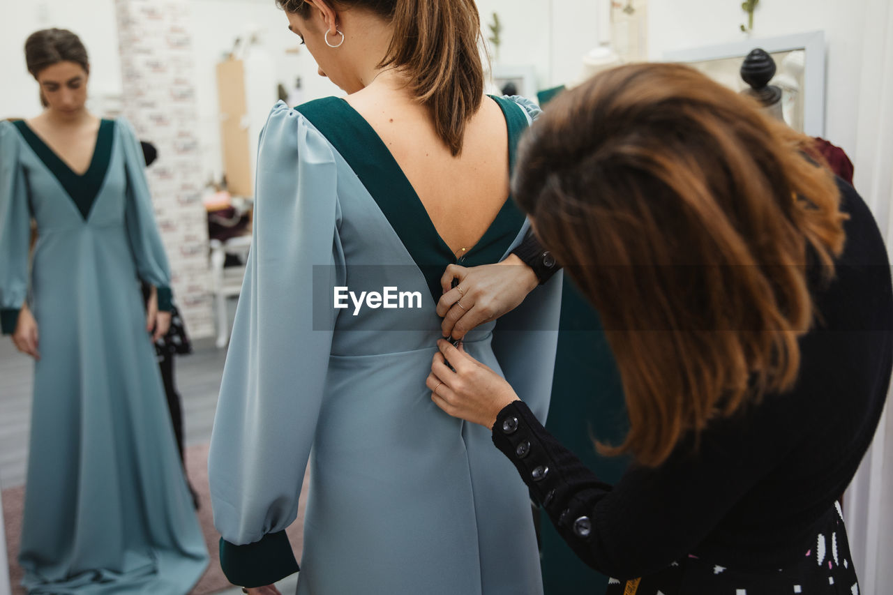 Unrecognizable dressmaker fitting custom gown on back of female client while working in professional workshop