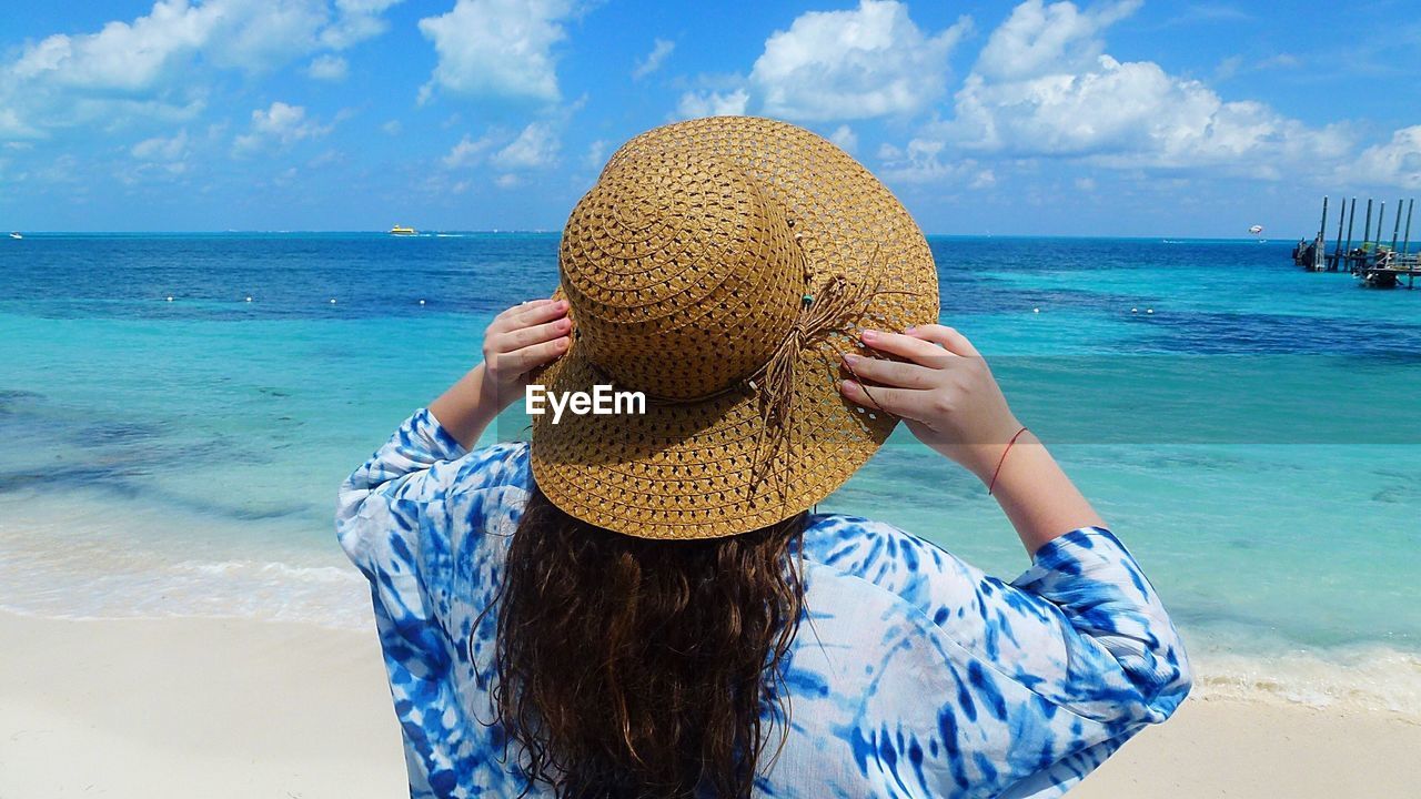 Rear view of woman in hat standing at beach against sky