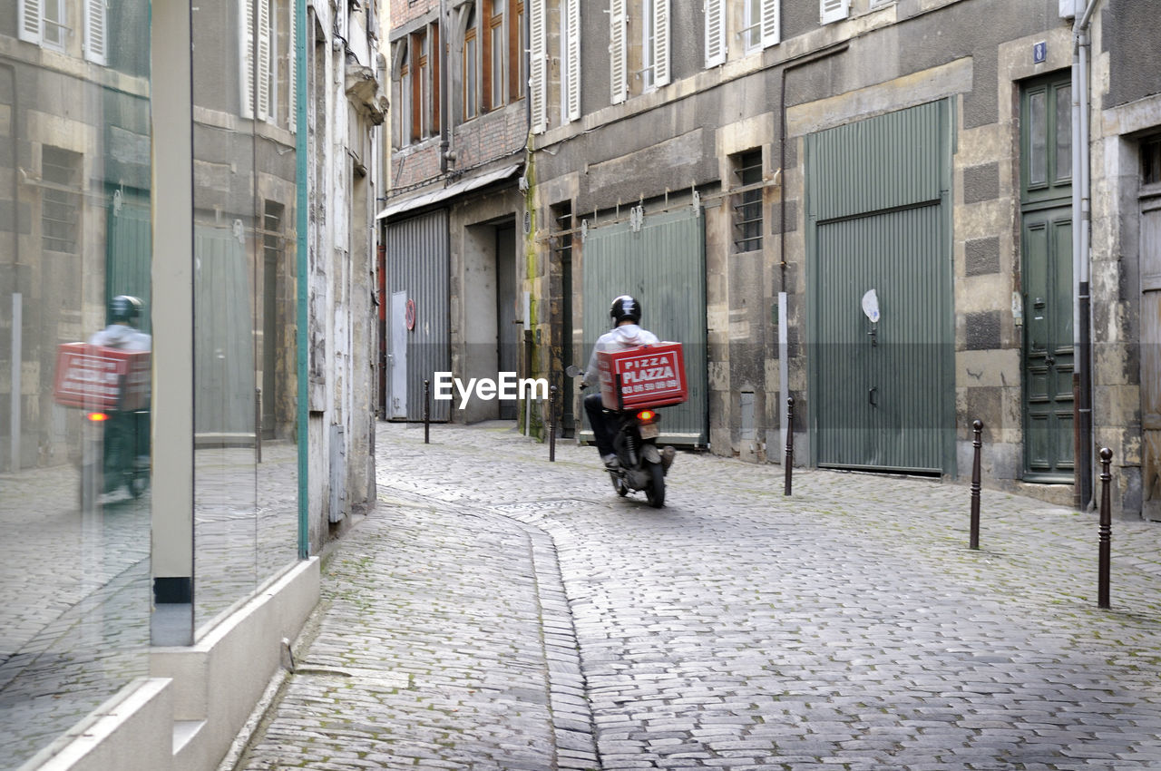 REAR VIEW OF MAN STANDING ON STREET AMIDST BUILDINGS