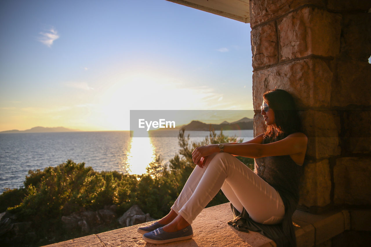 Smiling woman sitting on retaining wall by sea against sky during sunset