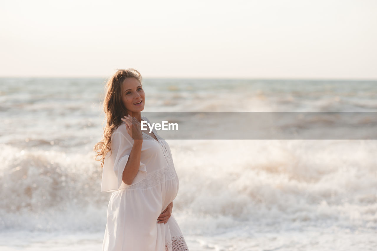 Beautiful laughing pregnant woman wear white dress posing over sea at background closeup.