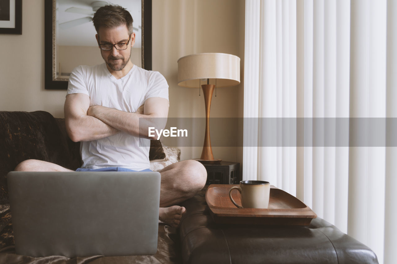 Man with arms crossed watching video on computer in warm morning light.