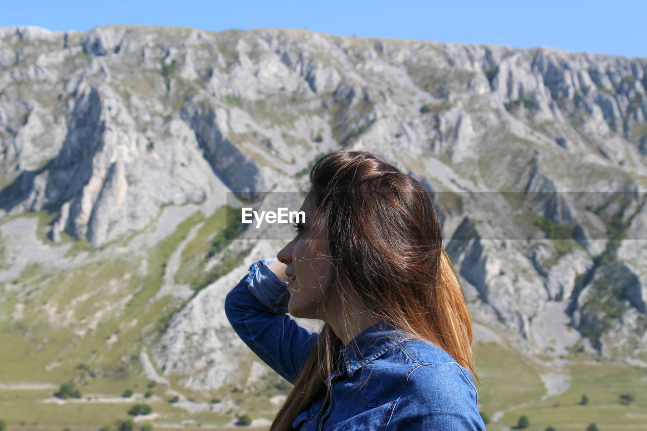 Side view of woman with hand in hair standing against mountains