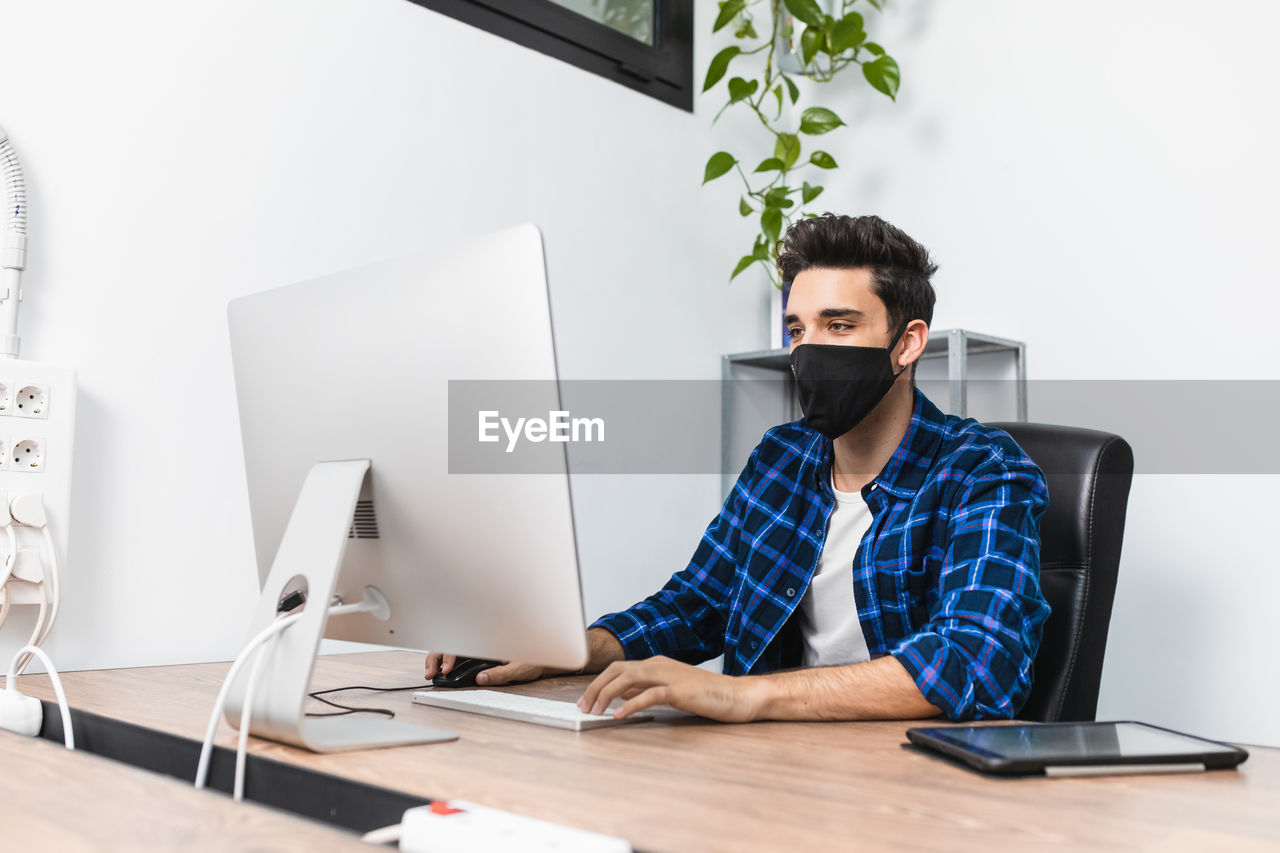 Unrecognizable young stylish male entrepreneur in face mask working on desktop computer in modern workspace