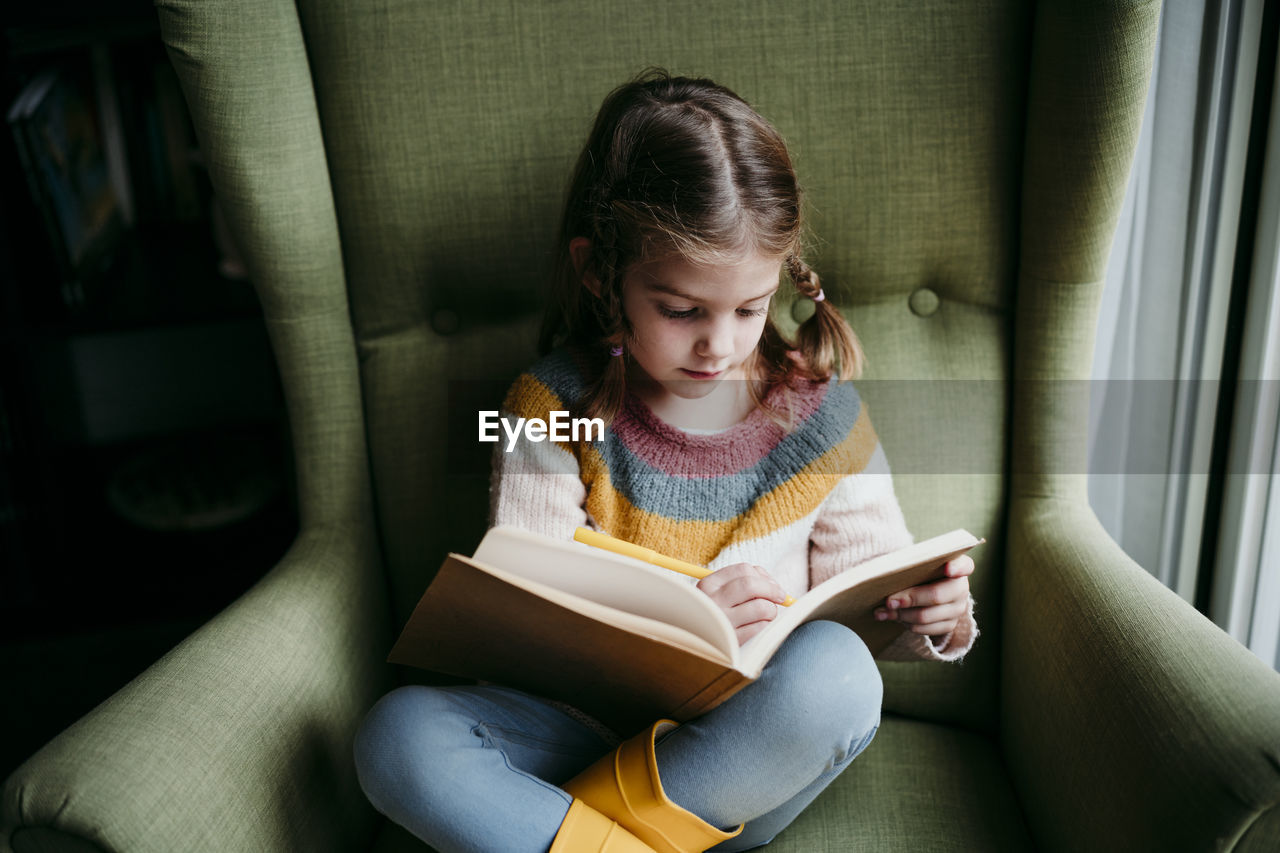 Cute girl writing in book while sitting on chair at home