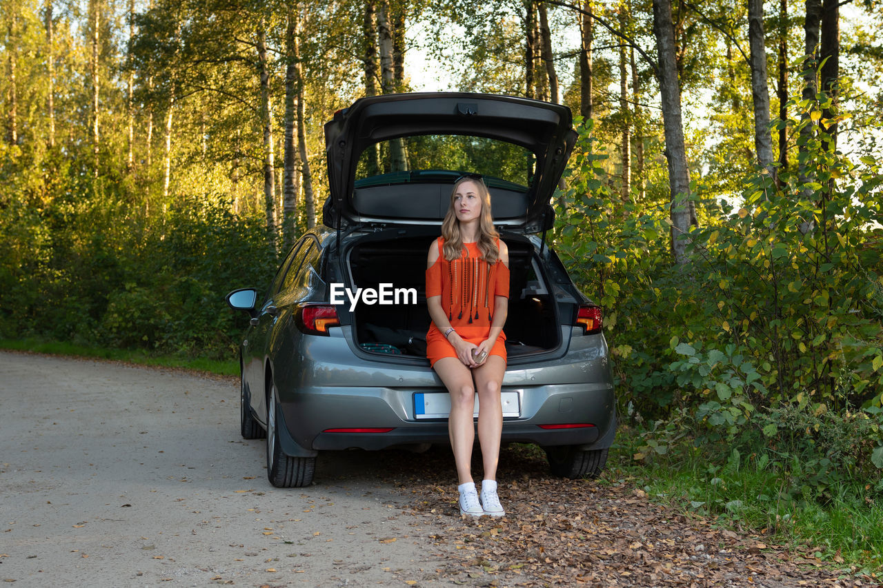 Woman sitting on car against trees in forest