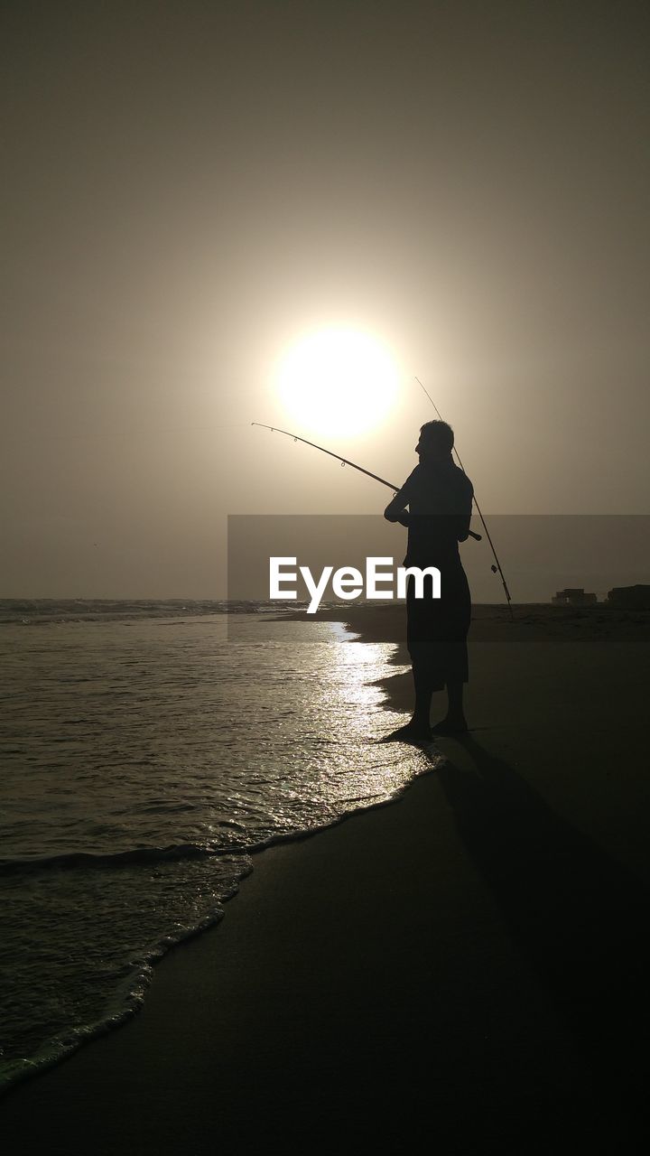 SILHOUETTE MAN STANDING AT SEA SHORE AGAINST SKY DURING SUNSET