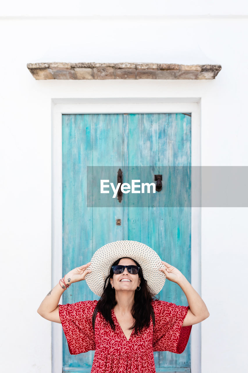 Female traveler in trendy apparel with arms holding hat and sunglasses near aged building in greece