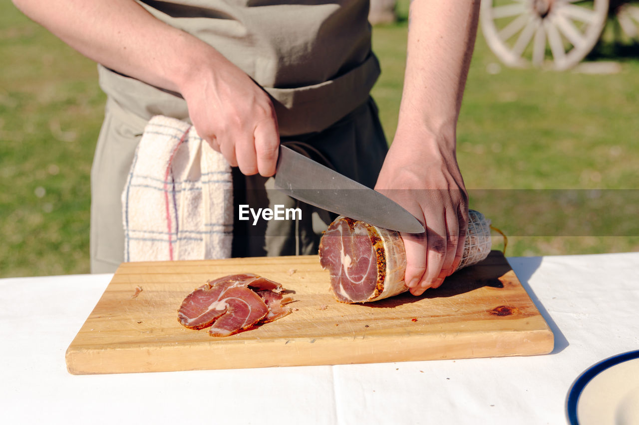 Unrecognizable crop cook standing at table in countryside and cutting delicious bresaola on wooden cutting board