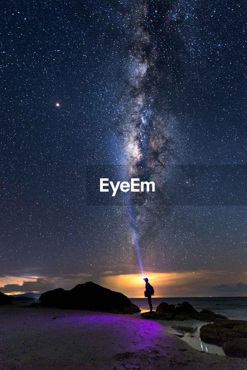 Silhouette person flashing light while standing on beach against sky at night