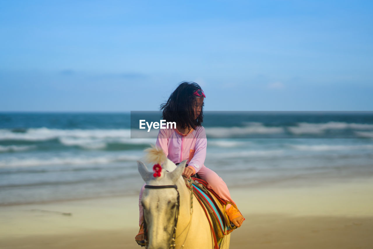 Rear view of girl riding on horse ft at beach against sky