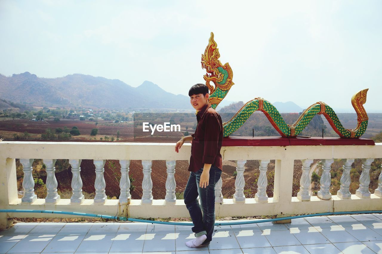 FULL LENGTH OF YOUNG MAN STANDING AGAINST RAILING AGAINST SKY