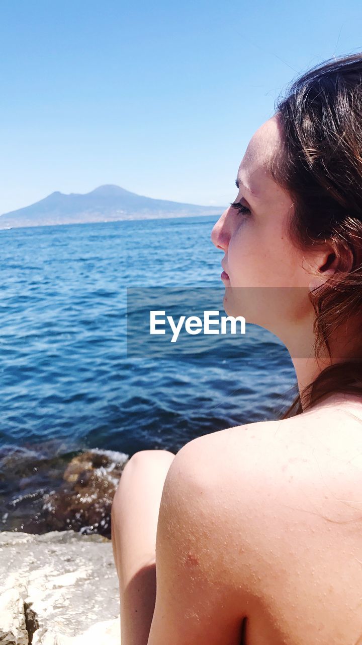 Rear of beautiful woman looking away while sitting by sea against clear sky during sunny day