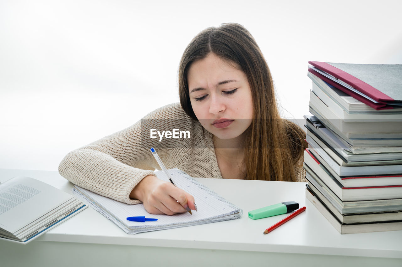 Worried teenage girl writing on book against white background